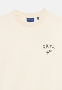 Helles beige Sweatshirt aus weichem Stoff, mit Rundhalsausschnitt und schwarzem Drucktext "QRTR 4TH" auf der linken Brust.