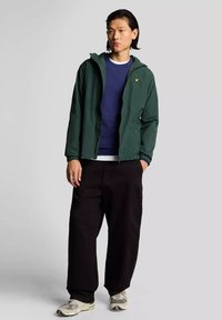 Giacca leggera verde con zip frontale, indossata sopra un maglione blu scuro, abbinata a pantaloni larghi neri e sneakers grigie.