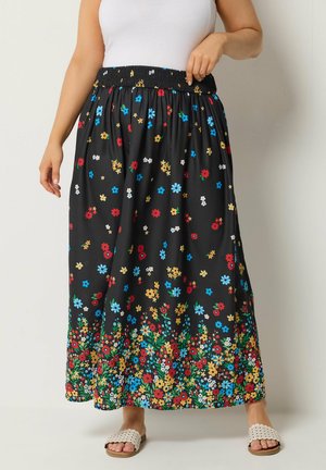 Kvinde iført sort maxinederdel med farverigt blomsterprint og elastisk talje, stylet med hvide hæklede slip-on sandaler.