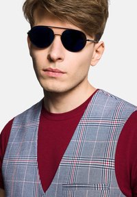 Czarne okulary przeciwsłoneczne aviator, szary kamizelka w kratę nałożona na bordową koszulkę z kieszonką i głębokim dekoltem w kształcie litery V. Gładkie faktury i kontrastowe kolory.