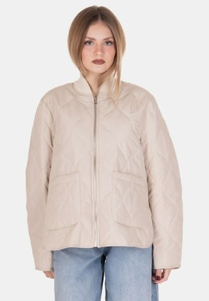 Giovane donna con capelli biondi fino alle spalle, che indossa una giacca beige trapuntata con zip e jeans azzurri chiari, in piedi su uno sfondo bianco.