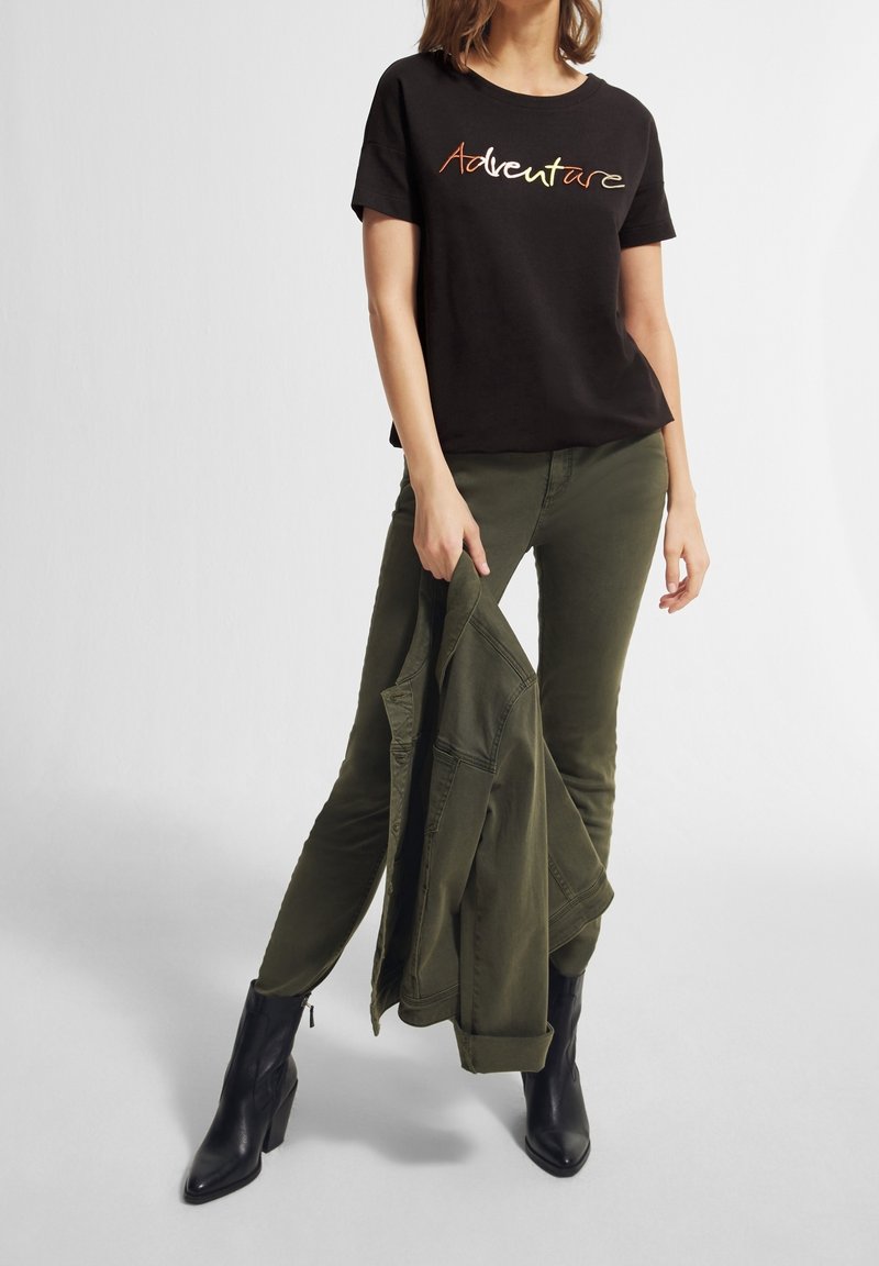 T-shirt nera a maniche corte con la scritta "Avventura" in lettere multicolori, abbinata a pantaloni verde oliva. Il modello tiene in mano una giacca verde. Stivali neri alla caviglia.