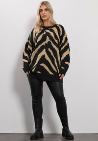 Kvinna i en överdimensionerad svart och beige zebra-mönstrad tröja, svarta fusk-läder leggings och svarta kraftiga stövlar, som står mot en enkel vägg.