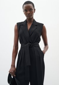 Combinaison sans manches noire avec un col ajusté, une taille ceinturée, un pantalon plissé et des poches latérales, confectionnée dans un tissu léger.