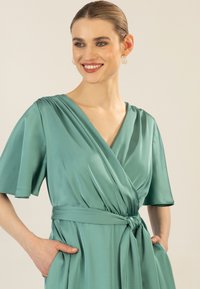 Un vestito avvolgente in satin teal con maniche corte a volant, una cintura da annodare e tasche laterali. Il tessuto ha una trama morbida e una leggera lucentezza.