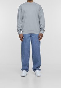 Felpa grigia chiara con polsini e orlo a coste, abbinata a jeans blu a taglio largo e sneakers bianche. Design di abbigliamento semplice e casual.