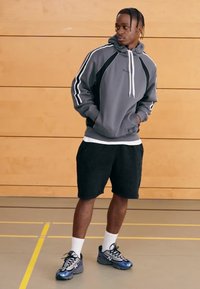 Grå huvtröja med vita detaljer, svarta shorts och blå/svarta träningsskor. Modellen står i ett gym med en vägg av trä.