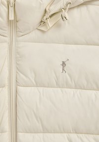 Kremfarvet quiltet jakke med lynlås, justerbare hættesnore og en lille broderet figur med en golfkølle på brystet.