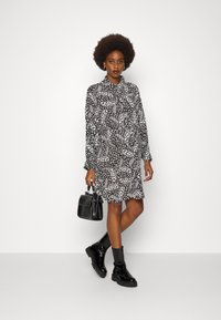 Vestido camiseiro com padrão preto e branco, mangas longas, colarinho e corte solto, combinado com botas cano curto pretas e uma pequena bolsa.