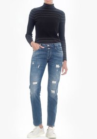 Jersey negro de cuello alto de canalé y jeans azul desgastados con parches azul claro. La modelo lleva zapatillas blancas y tiene las manos en los bolsillos.