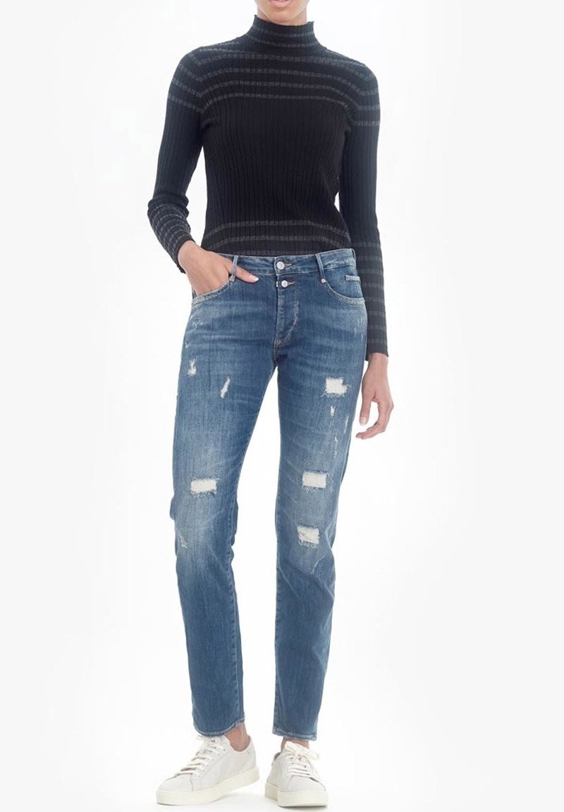 Jersey negro de cuello alto de canalé y jeans azul desgastados con parches azul claro. La modelo lleva zapatillas blancas y tiene las manos en los bolsillos.