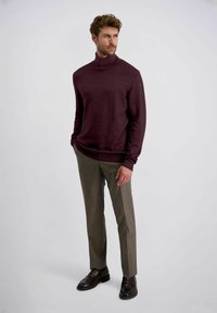 Man die staat met zijn handen in zijn zakken, gekleed in een bordeauxrode coltrui, grijze broek en donkerbruine leren schoenen.