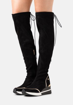 Czarne kozaki za kolano na koturnie z bocznym suwakiem i wiązaniem z tyłu, noszone na gołych nogach na jasnym, jednolitym tle.