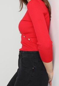 Czerwony prążkowany top z długim rękawem, zakrzywionym dekoltem i dwoma ozdobnymi metalowymi guzikami z boku, w połączeniu z wysokim stanem czarnych dżinsów.