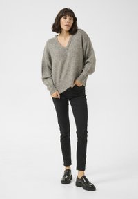 Hellgrauer, oversized Strickpullover mit V-Ausschnitt und geripptem Saum, kombiniert mit schwarzen Slim-Fit-Hosen und schwarzen Slippern.