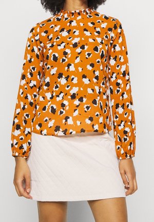 Kvinde iført orange langærmet bluse med sort og hvidt blomstermønster og lysefarvet quiltet miniskørt.