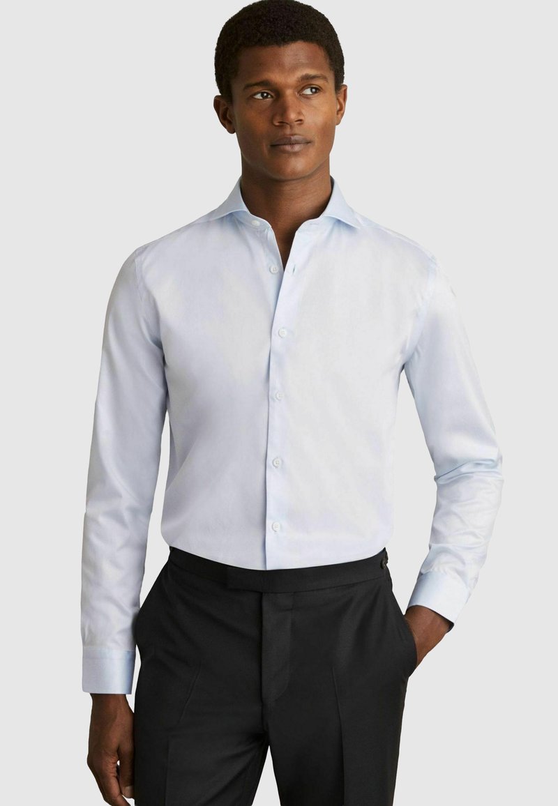 Chemise légère bleu clair avec un col classique, des poignets et un boutonnage à l'avant. Fabriquée en tissu lisse, portée avec un pantalon noir.