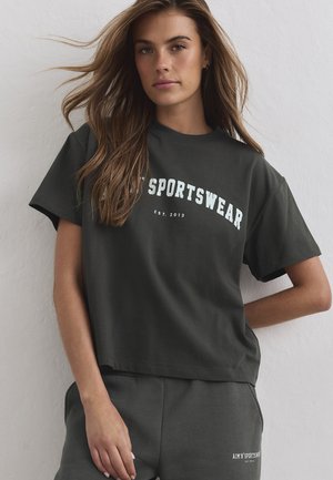 Femme aux longs cheveux bruns portant un t-shirt gris foncé et un short assorti avec un texte blanc de vêtements de sport.