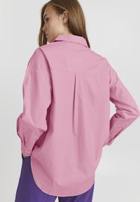 Rosa Langarmshirt mit Rückenfalten, kragenförmigem Halsauschnitt und geknöpften Manschetten. Glatte Textur, lockere Passform, kombiniert mit lila Hosen.