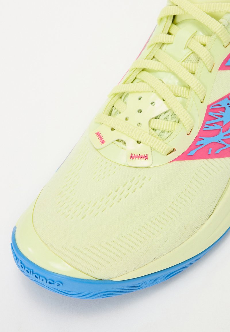 Zapatilla deportiva de color amarillo claro con una parte superior texturizada, acentos rosas y suela azul. Cuenta con un sistema de cordones seguro y paneles perforados.