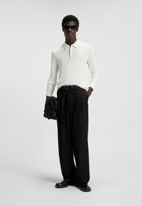 Polo shirt bianco a maniche lunghe con colletto, abbinato a pantaloni neri a gamba larga. Il modello tiene una giacca nera ed indossa occhiali da sole scuri.