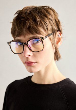 Lunettes écaille de tortue avec de grandes montures rectangulaires, dotées de verres réfléchissants et de branches fines, portées sur un fond neutre.