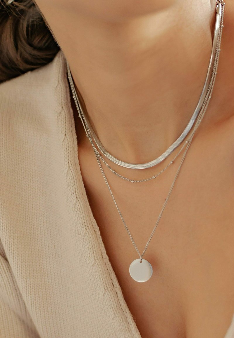 Collane a strati in argento con un pendente rotondo e piatto, stili di catena alternati e trame varie, indossate sopra un top in maglia beige.