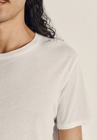 Bela bombažna majica s kratkimi rokavi in okroglim izrezom. Mehka tekstura in sproščen kroj, z minimalnimi šivi in brez vzorcev.