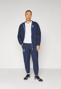 Marineblå windbreaker med lyseblå detaljer og logo, parret med matchende joggers og sorte sportssko. Har en lynlås og elastiske manchetter.
