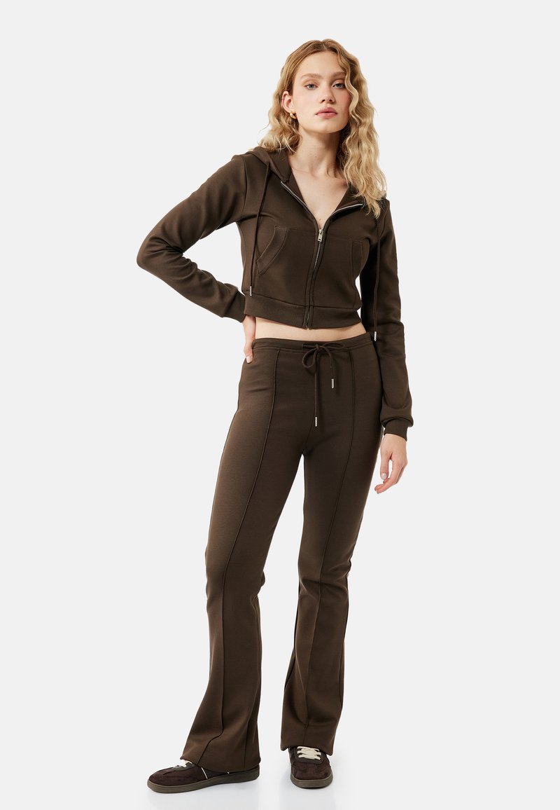 Jeune femme aux cheveux blonds ondulés portant un sweat à capuche court zippé marron foncé et un pantalon évasé assorti avec des baskets.