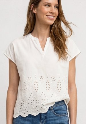 Frau lächelnd, trägt eine weiße kurzärmelige Bluse mit Lochmuster und gewelltem Saum, kombiniert mit blauen Jeans.