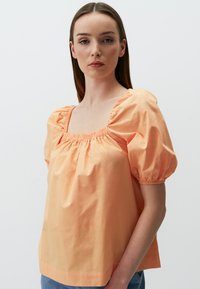 Blouse à manches bouffantes couleur pêche avec un décolleté carré et des détails froncés. Fabriquée en tissu léger et lisse.