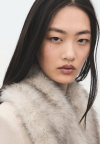 Primer plano de una persona que lleva un collar de piel suave de color gris claro. El fondo es neutro, lo que resalta la textura y el color del collar.