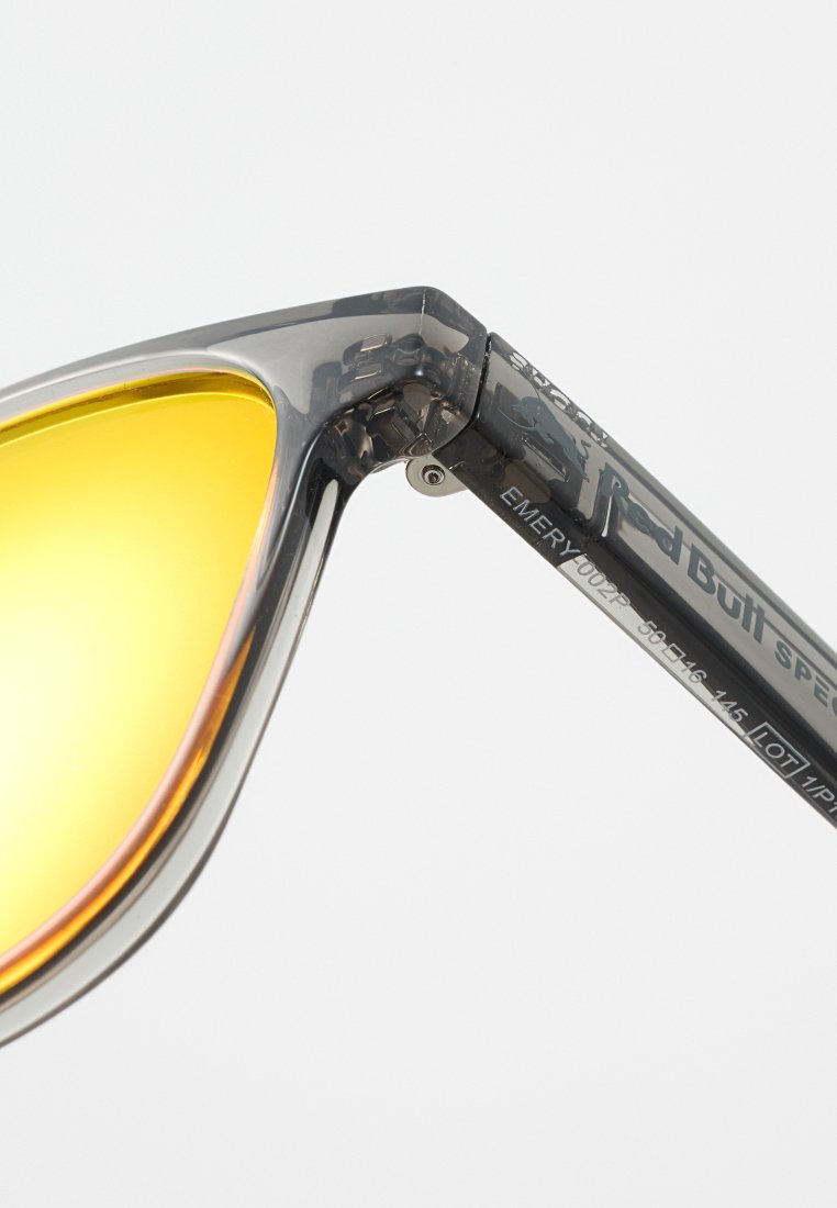 Okulary przeciwsłoneczne z przezroczystą szarym oprawką, soczewkami w żółtym odcieniu i srebrnymi elementami. Wyraźne logo na ramieniu, model EMERY 1002P.