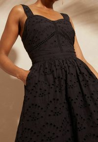 Žena ve bezramenných černých šatech s květinovými výřezy a s rukou v kapse šatů, na béžovém pozadí.