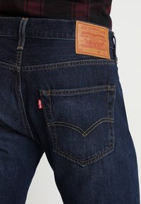 Vaqueros de mezclilla azul oscuro con costuras amarillas contrastantes, una etiqueta de cuero en la cintura y un sutil diseño de bolsillo curvado.