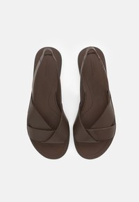 Pāris tumši brūnu ādas sandales ar platām krustojošām siksnām un plakanām zolēm uz baltas fona.