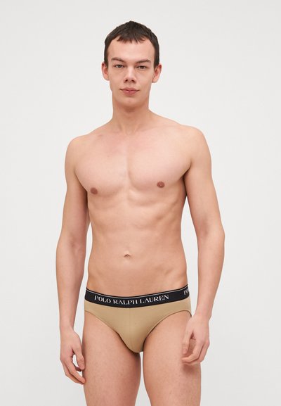 Μπεζ βαμβακερό σλιπ με μαύρη λωρίδα με επωνυμία. Το σλιπ έχει λεία υφή και εφαρμοστή σχεδίαση, αναδεικνύοντας τη μέση και τους γοφούς.