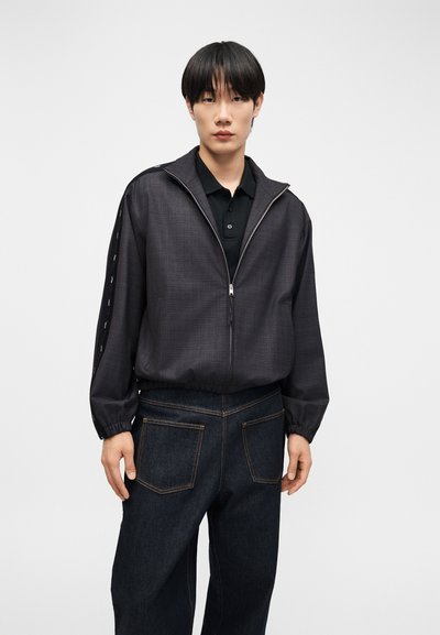 Tânăr purtând o jachetă închisă la culoare cu fermoar peste un tricou polo negru și blugi închiși la culoare, stând în picioare pe un fundal simplu, deschis la culoare.