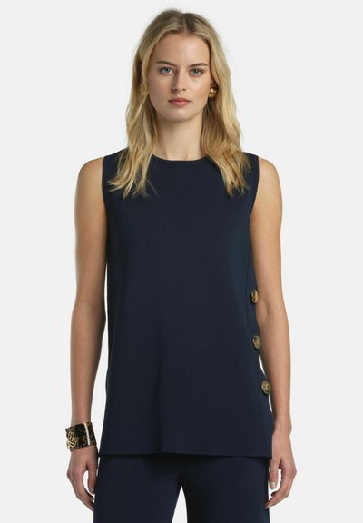 Femme blonde portant un haut sans manches bleu marine avec trois boutons dorés d'un côté, un pantalon bleu marine assorti, des boucles d'oreilles dorées et un bracelet imprimé léopard.