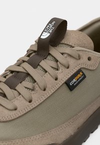 Beige sneaker featuring a textured upper made of Cordura fabric, with a padded tongue, brown laces, and branded heel tab.