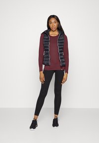 Svart puffer vest med glidelås over en burgunder langermet topp og svarte leggings. Svarte trening sneakers fullfører antrekket.