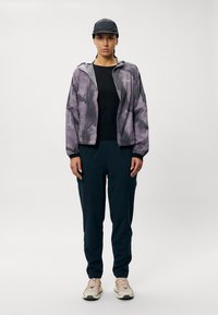 Lett, lilla og gråmønstret jakke med elastiske mansjetter, sammen med mørke joggebukser og beige joggesko med snøring.