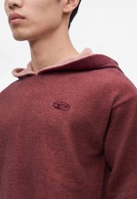 Jeune personne portant un sweat à capuche bordeaux avec un petit logo brodé sur la poitrine, visible du cou jusqu'au milieu du torse.