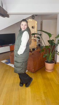 Colete acolchoado longo verde com gola alta, camisa branca de mangas compridas por baixo, combinado com calças pretas e sapatos pretos, ambientado numa sala aconchegante.