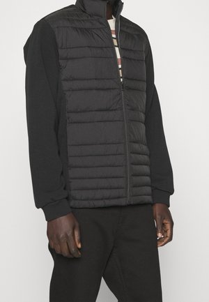 Hombre con chaleco negro acolchado con cremallera, sobre una camisa negra de manga larga y pantalones negros, de pie contra un fondo blanco.