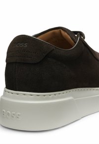 Brązowy zamszowy sneaker z białą gumową podeszwą, tonalnym przeszyciem i wytłoczonym logo "BOSS" na pięcie i boku. Zaokrąglony kształt noska.