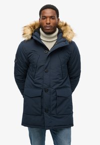 Giubbotto parka blu navy con cappuccio foderato in pelliccia sintetica, caratterizzato da chiusure a bottone, due tasche frontali e una calda texture trapuntata.