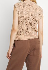 Stickad ärmlös tröja i ljusbeige med ett texturerat spetsmönster. Har en ribbad krage och fåll, kombinerad med bruna byxor.