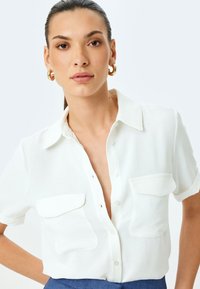 Witte kortekraagshirt met korte mouwen, voorzien van twee voorzakken en een knoopsluiting. Gemaakt van lichtgewicht stof met een subtiele textuur.